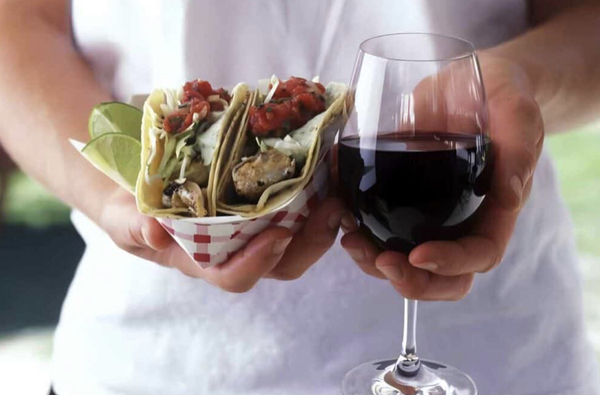 A close up of a woman's hands holding a glass of red wine in one hand and a plate of tacos in the other
